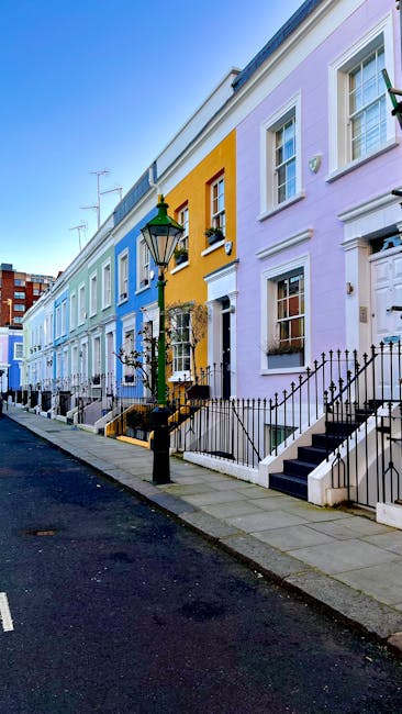 A row of colourful terraced houses in Hampstead Village, featuring pastel facades in shades of blue, yellow, and lavender. The houses have white window frames and black wrought iron railings alongside steps leading to front doors. A classic black street lamp is mounted on a post near the sidewalk, which is clean and free of debris. The street appears quiet, with minimal traffic, and bright daylight illuminates the scene, emphasizing the vibrant colors of the buildings. This image exemplifies the charming residential architecture typical of Hampstead, aligning with domestic cleaning themes highlighted by Hampstead Carpet Cleaners in their end of tenancy cleaning checklist. The scene suggests a well-maintained neighborhood, suitable for highlighting surface cleaning and home hygiene services.