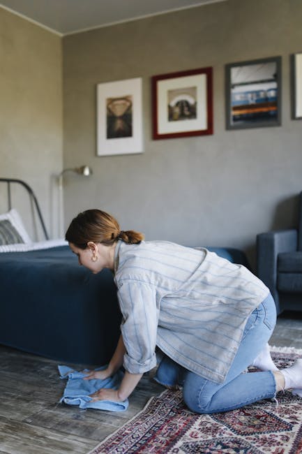 A woman kneeling on a patterned area rug, wearing a light blue striped shirt and jeans, is cleaning a wooden floor with a blue cloth in a well-lit living room. The room features a grey wall with three framed artworks hanging above a blue sofa and a matching armchair. The scene highlights domestic surface cleaning, emphasizing the importance of thorough and sanitised home maintenance, with Hampstead Carpet Cleaners specialising in deep cleaning services to maintain hygiene and cleanliness in residential spaces.