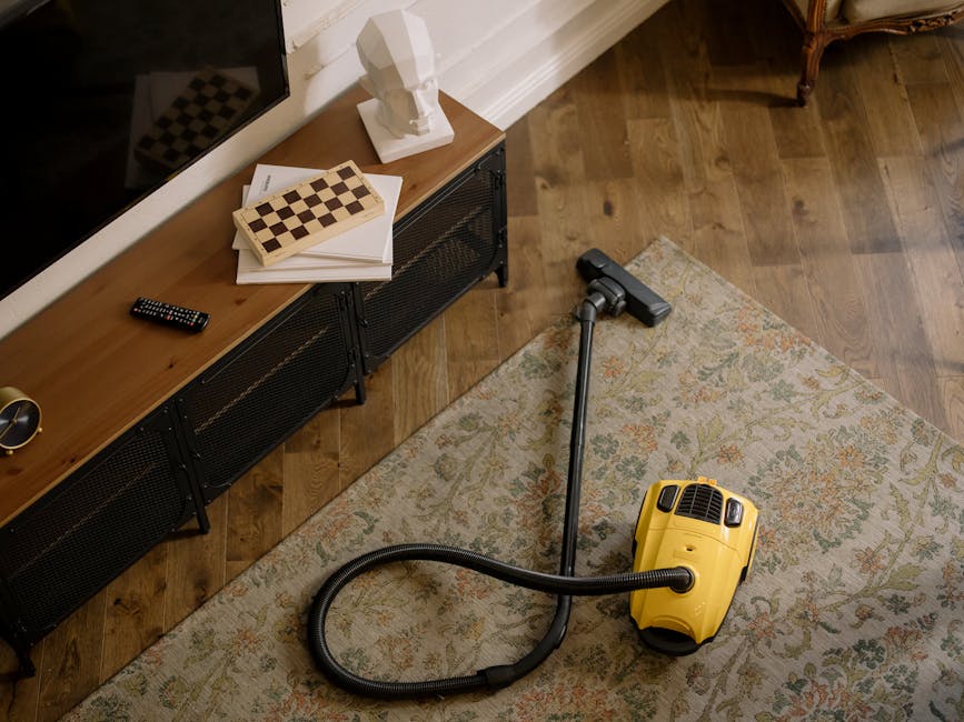 A woman kneeling on a patterned area rug, wearing a light blue striped shirt and jeans, is cleaning a wooden floor with a blue cloth in a well-lit living room. The room features a grey wall with three framed artworks hanging above a blue sofa and a matching armchair. The scene highlights domestic surface cleaning, emphasizing the importance of thorough and sanitised home maintenance, with Hampstead Carpet Cleaners specialising in deep cleaning services to maintain hygiene and cleanliness in residential spaces.