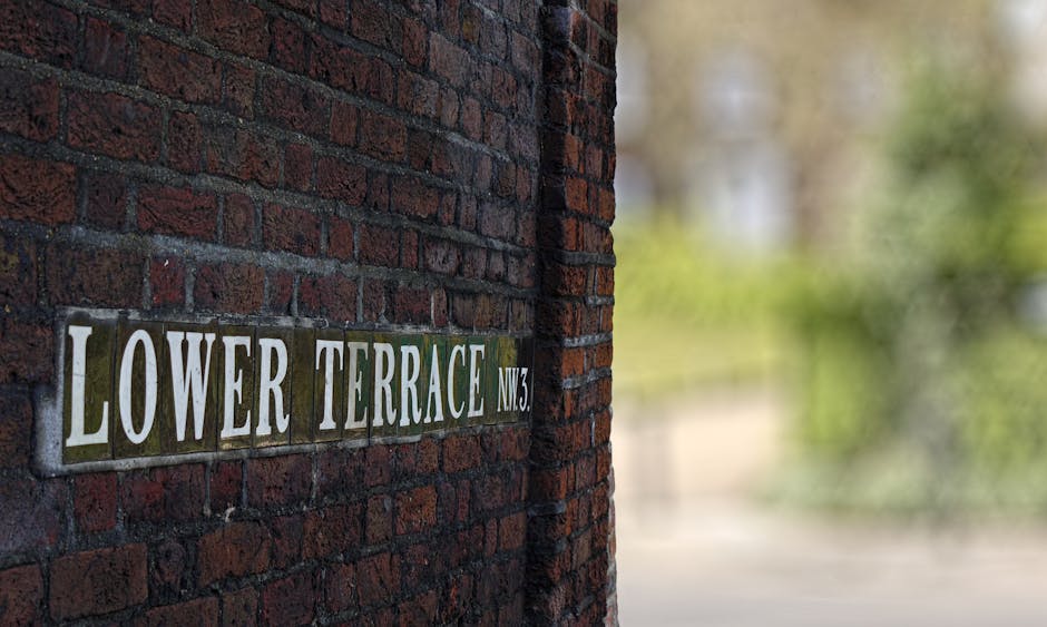 A brick wall with a street sign reading 'Lower Terrace NW3' attached to its surface. The sign has white capital letters on a dark background and is positioned on the left side of the image. The remaining brick wall appears to be weathered with a mix of dark red and brown bricks. In the background, blurred greenery is visible, indicating an outdoor setting with natural light illuminating the scene. This image emphasizes urban street signage and exterior brickwork, captured during daylight hours, showcasing a clean and well-maintained appearance in an outdoor environment. Hampstead Carpet Cleaners would focus on surface cleaning and maintenance of residential or commercial exteriors in similar settings.