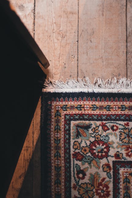 Close-up of a traditional patterned rug placed on a wooden floor near a black piece of furniture, such as a table or sofa, with the rug's edge featuring white fringe. The wooden flooring has a natural